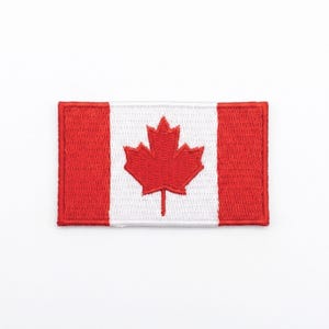 Puede incluir: Parche bordado con la bandera de Canadá. El diseño incluye una franja blanca central con una hoja de arce roja, flanqueada por dos franjas rojas verticales. El parche es rectangular y tiene un borde cosido.