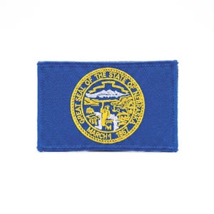 May include: A rectangular blue embroidered patch featuring the Great Seal of the State of Nebraska in yellow and white thread. The circular seal depicts a blacksmith, mountains, and a river, with the text Great Seal of the State of Nebraska March 1 1867.