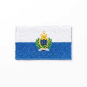 May include: Embroidered patch featuring the flag of San Marino. The rectangular patch has a white upper section and a blue lower section. The center displays the San Marino coat of arms, a gold crown over a shield with three towers, flanked by green laurel branches.