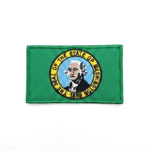 Könnte beinhalten: Ein rechteckiger bestickter Aufnäher mit der Flagge des Bundesstaates Washington. Das Design zeigt einen einfarbigen grünen Hintergrund mit einem zentralen gelben kreisförmigen Siegel, das ein Porträt von George Washington und den Text 1889 enthält.
