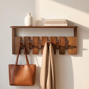 Puede incluir: Un perchero de pared de madera con una balda, ganchos y un bolso tote de cuero marrón. Una bufanda beige cuelga de uno de los ganchos. Un jarrón blanco y libros están en la balda. El perchero es de madera oscura.