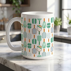 May include: White ceramic mug featuring a repeating pattern of pickleball paddles in shades of green, blue, and orange. The paddles have various designs, including stripes, diamonds, and solid colors. The mug has a large handle and is suitable for hot or cold beverages.