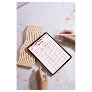 Op de afbeelding: Een digitale tablet met een "Client Booking Tracker" sjabloon, met een raster voor planning. De tablet wordt door handen vastgehouden, met een stylus in de buurt. Een lichtbruin, golvend houten decoratiestuk staat links.