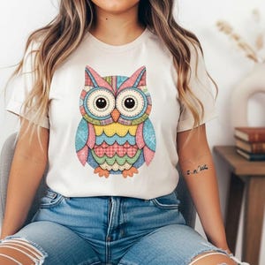Puede incluir: Una camiseta color crema con un diseño de búho colorido. El búho está formado por patrones de estilo patchwork en tonos de azul, rosa, amarillo y naranja. El búho tiene ojos grandes y redondos y está centrado en la camiseta.