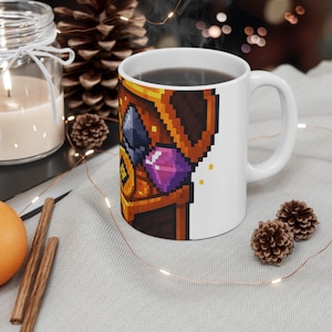 May include: White ceramic mug featuring a pixelated treasure chest design with gems. The mug is filled with a dark liquid and has a simple handle. The design includes shades of brown, gold, purple, and blue.
