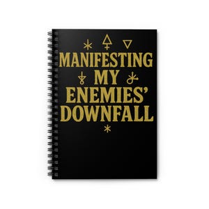 May include: Black spiral-bound notebook with gold lettering and symbols. The text reads "Manifesting My Enemies' Downfall." The notebook is closed and has a clean, minimalist design.