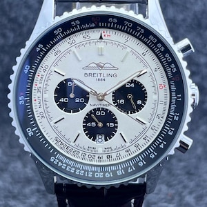 May include: A Breitling Navitimer 1884 watch with a silver dial, three black sub-dials, and a date window at the six o'clock position. The watch features a stainless steel case, a black leather strap, and a rotating slide rule bezel with markings.