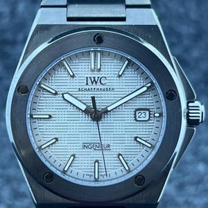 May include: A silver IWC Schaffhausen Ingenieur watch with a textured face and a date display at 3 o'clock. It has a stainless steel strap and a round bezel. The hands and hour markers are silver.