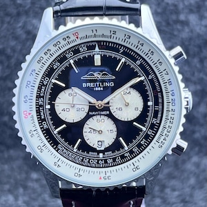 Könnte beinhalten: Eine Breitling Navitimer 1884 Armbanduhr mit schwarzem Zifferblatt, drei silbernen Hilfszifferblättern und einem schwarzen Lederarmband. Die Uhr verfügt über eine kreisförmige Rechenschieber-Lünette mit weißen Markierungen und silberne Zeiger.