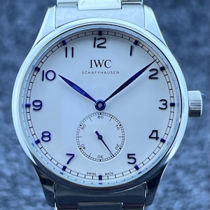May include: A stainless steel wristwatch with a white face and blue hands. The watch features the brand name "IWC SCHAFFHAUSEN" and a small seconds dial. The watch has a silver strap and a crown on the right side.