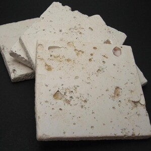 May include: Four square, off-white, textured coasters made of travertine stone. The coasters have a natural, rustic look with a variety of small holes and indentations.