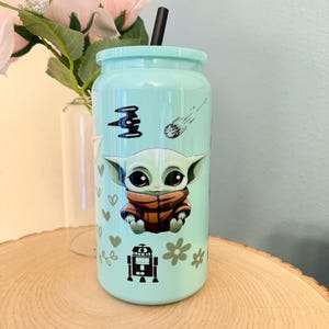May include: A light blue tumbler with a black straw, featuring a cartoon character design. The design includes a character in a brown coat, a spaceship, a shooting star, hearts, and flowers. The tumbler is on a wooden surface.