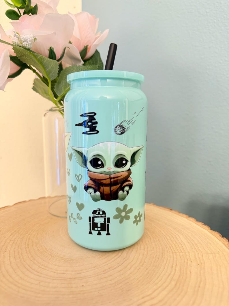 May include: A light blue tumbler with a black straw, featuring a cartoon character design. The design includes a character in a brown coat, a spaceship, a shooting star, hearts, and flowers. The tumbler is on a wooden surface.