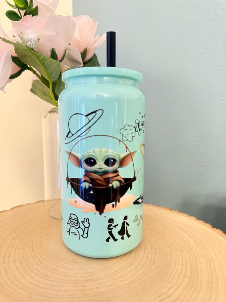 May include: A light teal tumbler with a black straw and lid. The tumbler features a cartoon image of a character from a popular science fiction franchise, along with other space-themed illustrations in black.