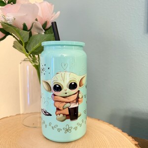 May include: A light blue tumbler with a cartoon image of a character holding a beverage. The tumbler has a black straw and a matching lid. Decorative elements include hearts and floral designs.