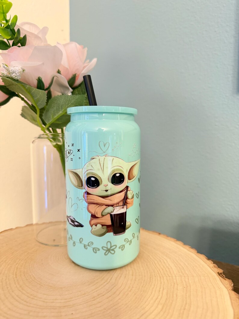 May include: A light blue tumbler with a cartoon image of a character holding a beverage. The tumbler has a black straw and a matching lid. Decorative elements include hearts and floral designs.
