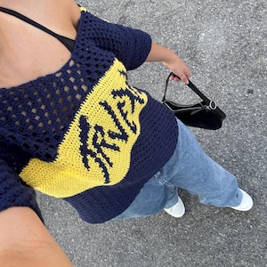 Puede incluir: Un top de ganchillo azul marino y amarillo con la palabra "IRVINE" en amarillo. El top se combina con vaqueros azules, un bolso negro y zapatillas blancas. El top tiene un cuello redondo y mangas cortas.