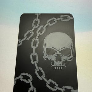 May include: A black rectangular card with a gray skull and chain design. The skull has detailed eye sockets and teeth. The chain is draped across the card, creating a gothic or edgy aesthetic.