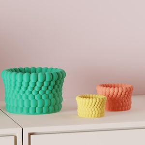 May include: Three decorative planters in shades of green, yellow, and orange sit on a white surface. The planters have a textured, bubble-like design. A stack of books is visible in the background.