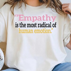 Può includere: Una t-shirt color crema con il testo "Empathy is the most radical of human emotion." La parola "Empathy" è stampata in rosa, la frase "is the most radical of" è in nero e "human emotion." è in giallo.