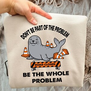 Puede incluir: Una sudadera beige con un gráfico de una foca gris rodeada de conos de tráfico naranjas y blancos y una barrera de carretera cerrada. El texto dice: DON'T BE PART OF THE PROBLEM BE THE WHOLE PROBLEM en letras mayúsculas negras.