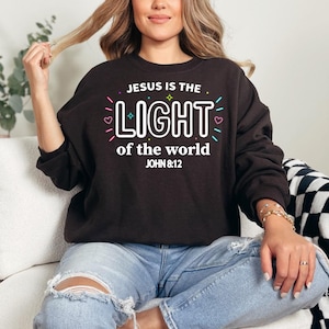 Könnte beinhalten: Ein schwarzes Langarm-Sweatshirt mit dem weißen Text "JESUS IS THE LIGHT of the world JOHN 8:12" mittig auf der Brust. Das Design enthält farbenfrohe grafische Akzente wie kleine Sterne, Herzen und Striche in Pink, Blau und Gelb.