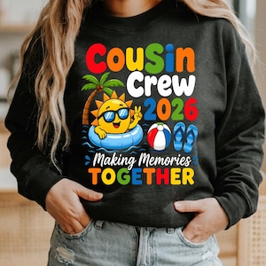 Może przedstawiać: Ciemnoszara bluza z kolorową grafiką z napisem "Cousin Crew 2026 Making Memories Together". Projekt zawiera kreskówkowe słońce w okularach przeciwsłonecznych, palmę, piłkę plażową i klapki, w letnim klimacie.
