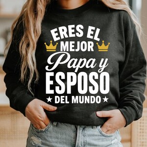 May include: A black long-sleeve sweatshirt featuring white text that reads ERES EL MEJOR Papa y ESPOSO DEL MUNDO, flanked by two gold-colored crown icons and two white stars, printed on the front center of the garment in a mix of fonts.