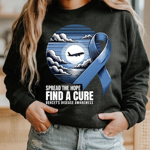 May include: Dark gray sweatshirt featuring a blue ribbon and a circular graphic with a plane, clouds, and a moon. The text reads "SPREAD THE HOPE FIND A CURE BEHCET'S DISEASE AWARENESS".