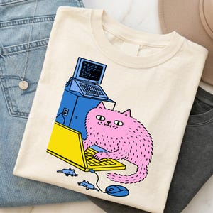 Könnte beinhalten: Cremefarbenes T-Shirt mit einer rosa, flauschigen Katze, die einen gelben Laptop benutzt. Die Katze sitzt vor einem blauen Computerturm mit einem Laptop-Bildschirm. Eine blaue Maus und zwei blaue Mäuse sind ebenfalls im Bild. Das Shirt liegt auf blauen Jeans und einem grauen Shirt.