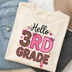 Könnte beinhalten: Ein cremefarbenes T-Shirt mit dem Text Hello 3rd Grade in Schwarz, Pink und Leopardenmuster. Das Design enthält pinke Schleifen, kleine pinke Herzen und goldene Sterne. Die Zahl 3 ist im Leopardenmuster, während die Buchstaben pink kariert sind.