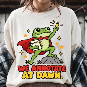 May include: A cream-colored sweatshirt featuring a green frog wearing a red cape and bow tie, holding a yellow highlighter and a notebook labeled NOTES. The text WE ANNOTATE AT DAWN is printed below the frog, surrounded by yellow star icons.