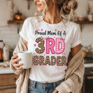 Könnte beinhalten: Ein weißes Kurzarm-T-Shirt mit dem schwarzen Text Proud Mom Of A 3RD GRADER. Die Zahl 3 ist in Leopardenmuster, die Buchstaben RD in rosa Karomuster und das Wort GRADER in Leopardenmuster gehalten, alles akzentuiert mit kleinen rosa Schleifen.