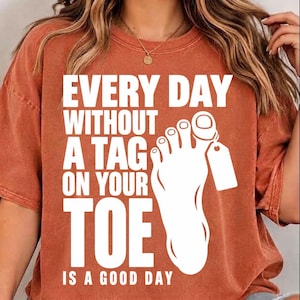 May include: A rust-colored short-sleeve t-shirt featuring white text that reads EVERY DAY WITHOUT A TAG ON YOUR TOE IS A GOOD DAY, accompanied by a white graphic of a human foot with a blank identification tag attached to the big toe.