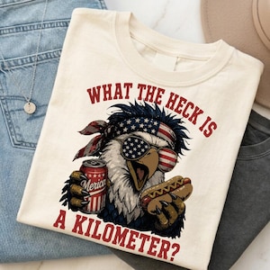 May include: A cream-coloured t-shirt featuring a graphic of a bald eagle wearing a stars and stripes bandana and sunglasses, holding a soda can and a hot dog. The text reads: WHAT THE HECK IS A KILOMETER? The design uses red, white, and blue colours.