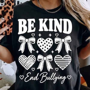 May include: A black short-sleeved t-shirt featuring white graphic text that reads BE KIND and End Bullying. The design includes three white bows and three hearts with different patterns, including polka dots, diagonal lines, and horizontal stripes.