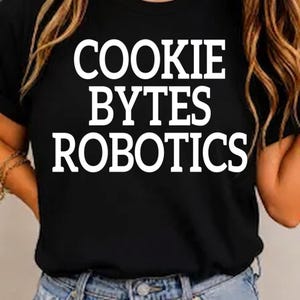 以下が含まれることがあります： 胸元に太字のサンセリフ体で白い文字「COOKIE BYTES ROBOTICS」がプリントされた黒の半袖Tシャツ。クルーネック仕様で、裾には青いデニムパンツが見えています。