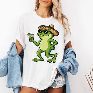 May include: A white t-shirt featuring a graphic illustration of a green frog wearing a colorful sombrero and black sunglasses, holding a margarita glass with a lime slice. The frog has a light green belly and is depicted in a standing pose.