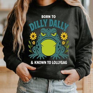 May include: A black long-sleeve sweatshirt featuring a graphic of a teal and lime green toad with yellow sunflowers. The text reads BORN TO DILLY DALLY & KNOWN TO LOLLYGAG in white and teal lettering centred on the front of the garment.