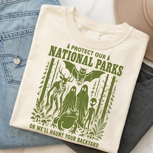 Może przedstawiać: Kremowy t-shirt z zielonym nadrukiem graficznym i napisem "PROTECT OUR NATIONAL PARKS OR WE'LL HAUNT YOUR BACKYARD." Wzór zawiera ilustracje lasu, nietoperza, stworzenia przypominającego jelenia, duchów, żaby oraz kosmity.