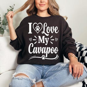 May include: A dark brown sweatshirt with the text "I Love My Cavapoo" in white. The design includes a heart with a paw print, hearts, and paw prints. The sweatshirt is worn with blue jeans.