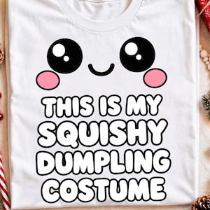 May include: A white t-shirt featuring a graphic of a smiling face with large black eyes and pink cheeks. Below the face, the text reads THIS IS MY SQUISHY DUMPLING COSTUME in bold, black-outlined white letters. The design is centered on the chest.