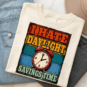 May include: A cream-coloured t-shirt featuring a graphic print with the text I HATE DAYLIGHT SAVINGS TIME in bold orange and yellow letters. The design includes a red twin-bell alarm clock set against horizontal stripes of orange, yellow, and blue.