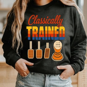 Może przedstawiać: Czarna bluza z długim rękawem z napisem Classically Trained w kolorze pomarańczowym oraz blokiem w stylu retro. Pod tekstem znajduje się graficzna ilustracja trzech pedałów samochodowych oraz gałki manualnej skrzyni biegów z pięcioma biegami.