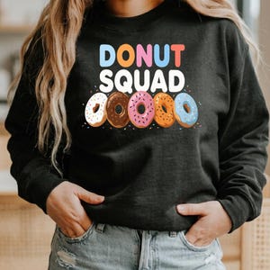 May include: A dark gray sweatshirt with the words "DONUT SQUAD" in colorful letters. Below the text are five donuts with different colors and toppings. The donuts are white, brown, pink, orange, and blue.