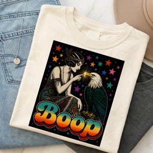 Könnte beinhalten: Ein cremefarbenes T-Shirt mit einem Grafikdruck einer Person im Flapper-Kleid, die den Schnabel eines Weißkopfseeadlers berührt. Das Design enthält bunte Sterne auf schwarzem Hintergrund und das Wort Boop in einer retro-farbigen Schriftart.