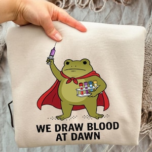 May include: A beige sweatshirt featuring a graphic of a green frog wearing a red cape, holding a syringe in one hand and a rack of colorful test tubes in the other. Below the illustration, the text reads: WE DRAW BLOOD AT DAWN.
