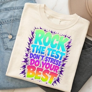 Può includere: T-shirt color crema con la scritta "ROCK THE TEST DON'T STRESS DO YOUR BEST" in un design colorato in stile graffiti. La maglietta è piegata e posizionata sopra un paio di jeans blu e una maglietta grigia.