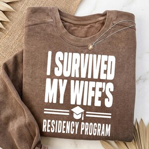 Könnte beinhalten: Ein braunes Sweatshirt mit Rundhalsausschnitt und Vintage-Waschung, das auf der Brust den weißen Aufdruck I SURVIVED MY WIFE'S RESIDENCY PROGRAM über einer Grafik einer Doktorhut-Kopfbedeckung und zwei horizontalen Linien zeigt.