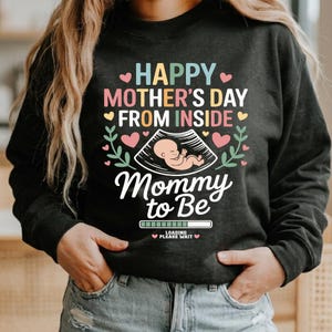 Può includere: Felpa grigio scuro con grafica colorata per la festa della mamma. Il design include il testo "HAPPY MOTHER'S DAY FROM INSIDE" e "Mommy to Be" con un'immagine ecografica di un bambino. Cuori rosa e foglie verdi decorano il design.